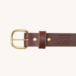Close-up of a brown leather work belt with a round solid brass buckle and two brass rivets, featuring the embossed Proven Hands logo on the strap.