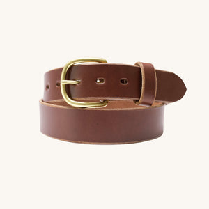 A close-up view of a brown full grain leather belt with a rounded solid brass buckle, coiled on a light background.